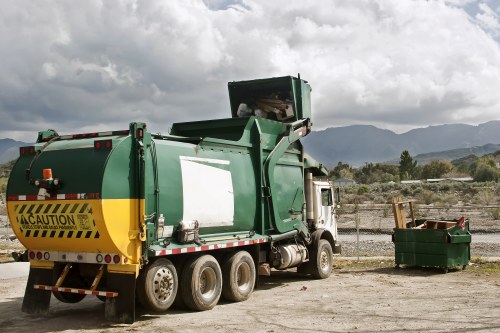 A large green waste collection truck with a white cab is parked on a gravel surface in an open outdoor area. The truck has a yellow caution sticker on the rear indicating safety, and the main body is designed for rubbish collection with a rear loading mechanism. To the right of the truck, there is a green skip bin filled with mixed waste materials, positioned next to a chain-link fence. The background features a landscape with distant hills and a cloudy sky, suggesting a rural or semi-industrial environment. The scene is typical of rubbish removal operations, with the truck and skip ready for waste disposal, fitting with services offered by Office Clearance Kings Cross for efficient rubbish clearance in the local area.