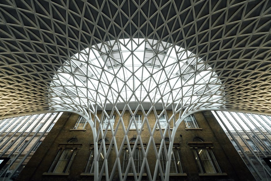 The interior of a modern train station featuring a large, vaulted ceiling with geometric lattice patterns made of metal, supporting a wide, dome-like glass roof that allows natural light to illuminate