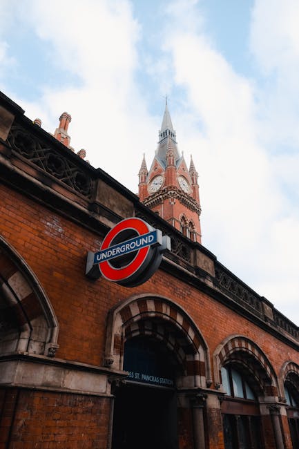 A close-up view of a red brick building with arched windows and a weathered surface, featuring a London Underground roundel sign prominently displayed in the foreground. Above the building, a section 