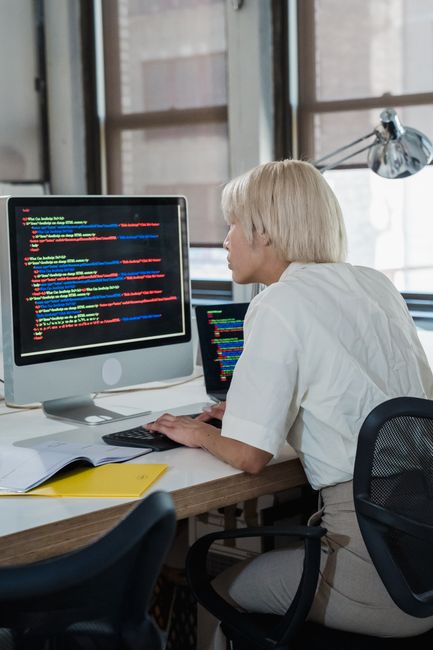 The image depicts a woman with short blonde hair working at a desk in a modern office environment. She is wearing a white shirt and is seated in front of a large desktop computer monitor displaying li